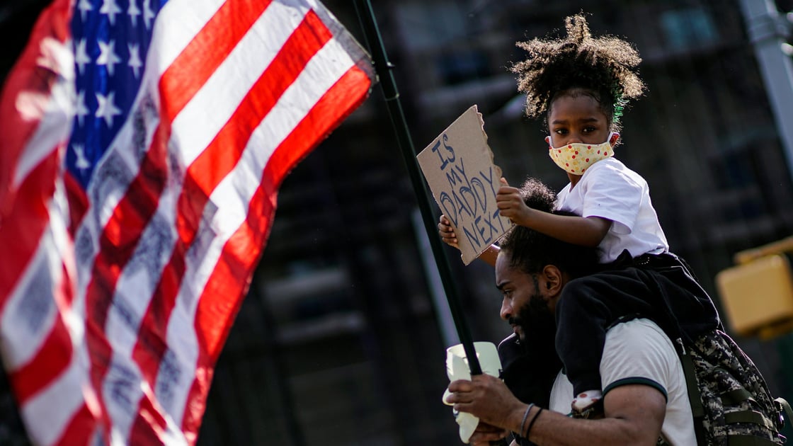 A child looks on with a sign while her father carries a U.S. flag during a protest against racial inequality