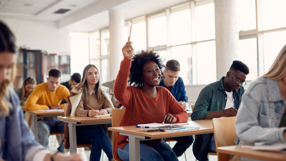 A high school student raises her hand in a classroom with other students.