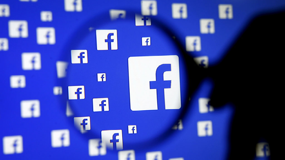 A man poses with a magnifier in front of a Facebook logo on display