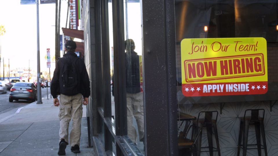 A pedestrian walks by a Now Hiring sign.