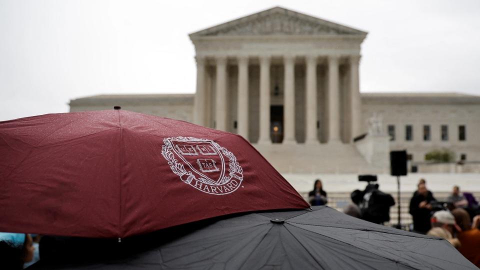 A person holds an umbrella with a Harvard logo print as demonstrators gather in support of affirmative action in front of the Supreme Court