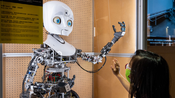 A young girl puts her hand up to a robot with a humanoid face and arm