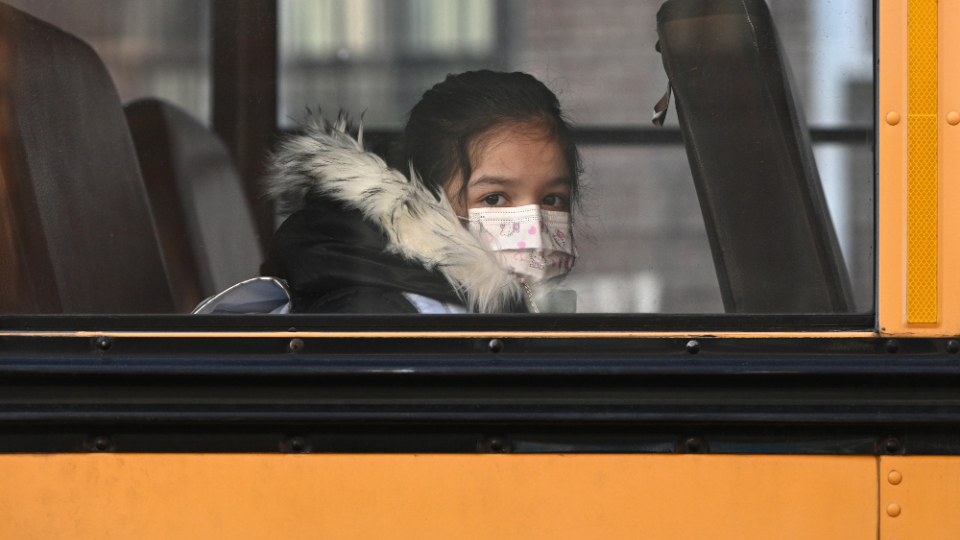 Child wears a mask on a school bus and looks out the window.