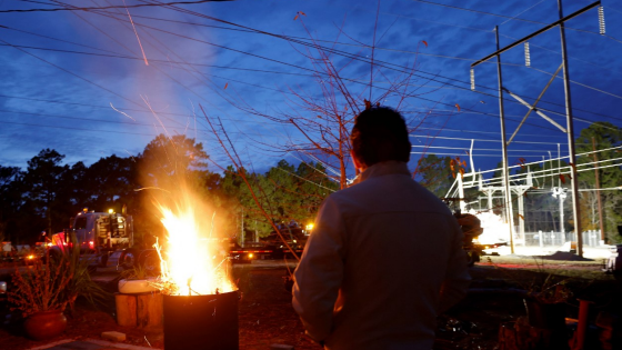 Gerardo Anicero warms himself in front of a makeshift fire as he watches Duke Energy personnel work to restore power at a crippled electrical substation