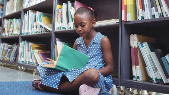 Girl reading book while sitting by shelf in library