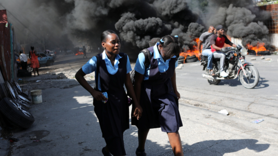 Girls wearing school uniforms walk past a burning street baricade during demonstrations in Haiti