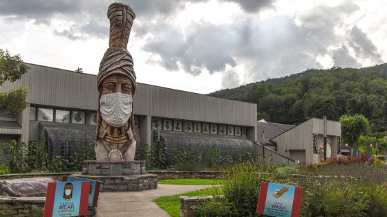 Large totem pole outside of the Museum of the Cherokee Indian with a face mask and signs for mask requirement