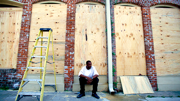 A young African-American man sits against a boarded up building. 