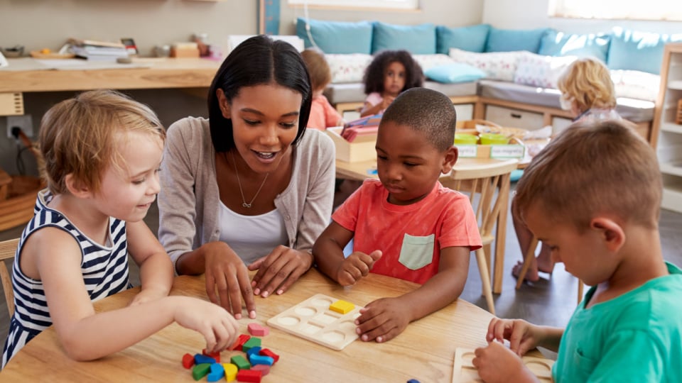 Preschool students work with teacher on block puzzle.