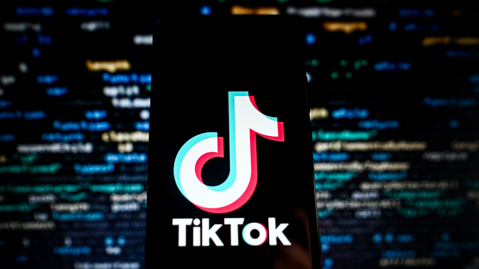 The TikTok logo is displayed on a smartphone with a screen showing programing codes on the background