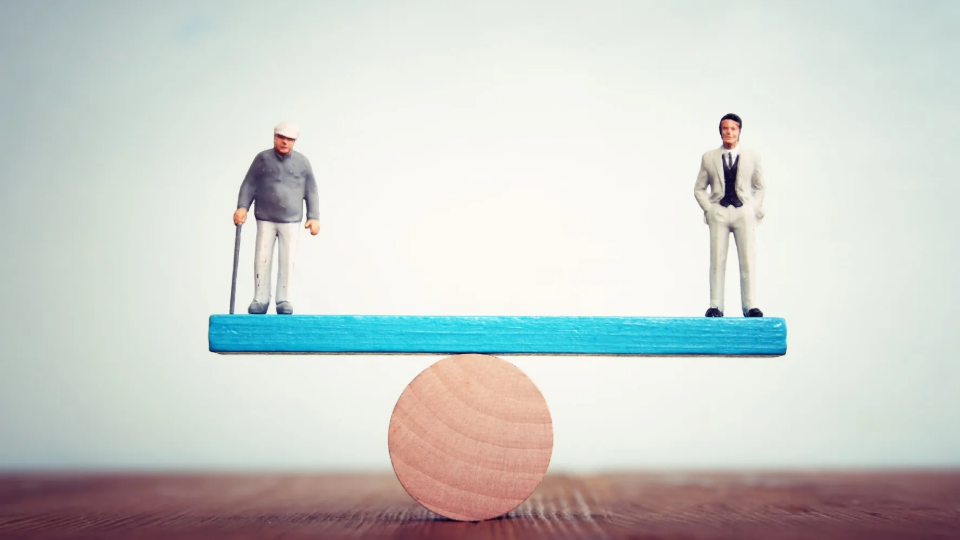 Toy representing elderly person and toy representing wealthy person are placed on a wooden scale