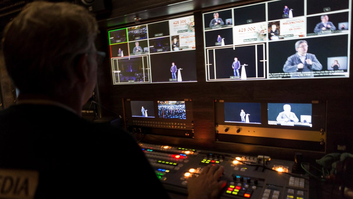 A man looks at a wall of computer screens to edit a video. 